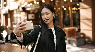 Happy young brunette lady in stylish black trench coat smiles sincerely, holds phone and takes selfie outside near street cafe.